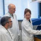 A seated male with grey hair and glasses, a standing bald male with glasses and a younger female with long brown hair tied in a ponytail stand in front of lab equipment. All three wear white lab coats and gloves. 