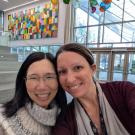 Two women smiling for a selfie in a bright atrium with colorful wall art and hanging glass orbs