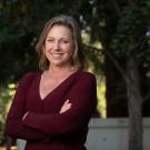 in an outdoor setting on a sunny day, a woman wearing a maroon sweater, has her arms crossed and is smiling at the camera.