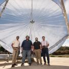 Four men stand outdoors in front of a large circular mirrored dish. 