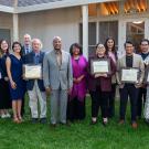 Group photo on lawn of diverse adults holding framed certificates outside a lighted building