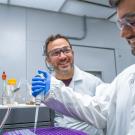 Two scientists in lab coats and goggles smiling while pipetting at a lab bench