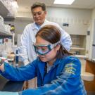 Two scientists in lab: woman pipetting while man observes, wearing goggles and gloves.