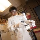 Undergraduate researcher wears safety goggles and gloves pours red liquid into a spray bottle labeled “70% EtOH Flammable” inside a laboratory workspace with shelves and equipment in the background.