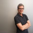 Smiling middle-aged man with glasses, arms crossed against a light gray wall.