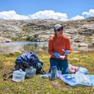 Person in orange jacket kneeling with camping gear beside a small alpine lake under blue sky