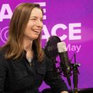 Smiling woman podcast host speaking into a microphone in a purple studio photo