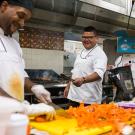 Two chefs are seen laughing while cooking inside an industrial kitchen. Chef Freddie Bitsoie is on the far right, facing the camera, in front of the grill.