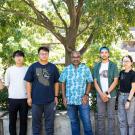 A group of seven people, casually dressed, outdoors under a tree, facing the camera