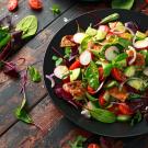 Mixed salad in dark bowl—greens, cherry tomatoes, radish slices and avocado on rustic wood