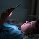 Girl lying in dark bedroom, face glowing from smartphone screen. UC Davis researchers have created a new public database that tracks bills regulating digital media for minors. (Adobe Stock)