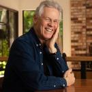Smiling man in blue shirt leaning on hand at wooden table in kitchen