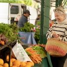 Woman holding woven basket chats with vendor at farmers market stall of carrots and greens