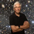 Older man with gray hair and glasses, arms crossed, smiling against starry galaxy backdrop