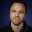 Smiling man with short dark hair and a beard, against a dark blue background.