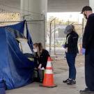 Three people stand outside of a tent under a freeway overpass