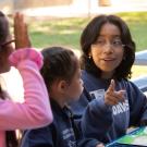 A female university student points at a child raising her hand during a reading lesson at an outdoor picnic table.