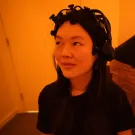 Woman with a specially designed headset in orange-lit room; man at laptop behind (photo)