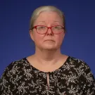 Headshot portrait of a person gray hair, red glasses, floral blouse against blue background