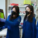 Three dark-haired women in dark blue lab coats look at a computer monitor to the left. 