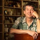Smiling man in patterned shirt leaning on a chair in warm-lit book-filled room