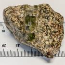 Roughly triangular pale off-white rock with brown and black flecks and a band green crystal running vertically across it. A ruler shows that it is about 50 millimeters across. 