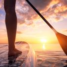 Legs shown of person on paddleboard in water toward sun