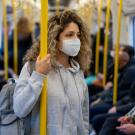 A young woman with long curly brown hair wearing a grey hoodie and face mask while riding light rail. Other passengers in the background also wear masks. 