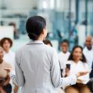 Woman with back to camera stands facing seated group of people listening to her