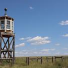 The guard tower at Amache National Historic Site, a Japanese Internment Camp in Colorado, rises high above the green grass around it on a bright, sunny day.