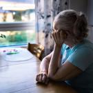 Older woman in blue shirt sitting at kitchen table, gazing pensively out window