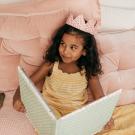 Young dark-complected girl with crown on head in pink-decorated bed looking at oversized book
