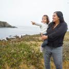 Woman in black shirt holds toddler daughter who is pointing at something. Ocean in background.