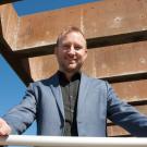 A man in a blue blazer stands confidently on a balcony under a clear blue sky.