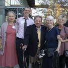 Six smiling adults outdoors, framed by a building a trees, one with a cane, one with a walker