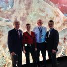 Four people in business attire face the camera. Behind them is a colorful, brightly lit wall display. 