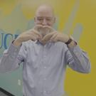 Robert Penman smiles and forms a heart with his hands in front of a colorful blue and yellow UC Davis mural. 