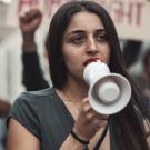 An Iranian female protestor holds a megaphone to her mouth. Behind her are blurred out protestors holding signs.