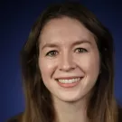 A person with long brown hair, wearing a dark green top, against a blue background.