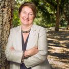 Smiling woman in beige blazer leaning against a tree in a sunlit park, portrait photo