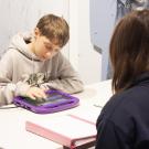 Boy focused on tablet at table, adult seated opposite observing.