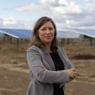 Professor Marina Leite in gray blazer, arms crossed, stands before rows of solar panels under cloudy sky