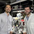 Jeremy Munday and Micah Karahadian smile beside equipmental equipmentin a lab, one wearing a white lab coat.