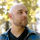 A bald man with a beard stands outdoors, wearing a plaid shirt, looking thoughtfully to the side.