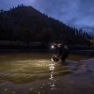 Dusk photo: person kneeling at riverbank, headlamp beam illuminating water