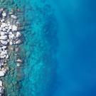 Aerial view of rocky shoreline with green bushes and clear turquoise-to-deep-blue water.