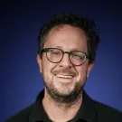 Smiling person in glasses, scruffy beard, black shirt — studio headshot, blue gradient background