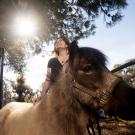 Sunlit photograph of smiling Rebecca Calisi Rodriguez standing behind a miniature horse, with trees and blue sky behind.