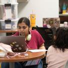 A young woman works on a laptop at a table, while a child sits nearby, looking away.
