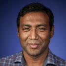 Smiling man with dark hair in a checked shirt against a blue background.